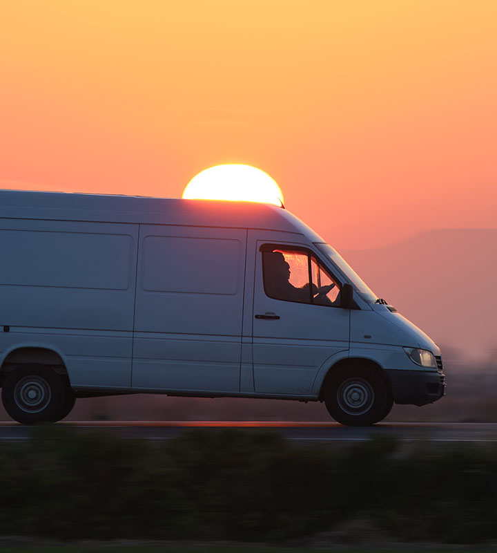 A white cargo van travels along a highway, silhouetted against a large, glowing orange sun setting behind the vehicle. The bright sunset illuminates the horizon during this evening transit.